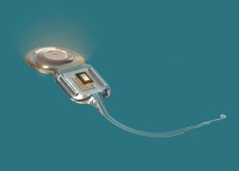 Close-up of a cochlear implant component, showing the processor unit with a curved wire/lead extending to the right on a teal background (medical device).