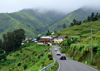 Winding mountain road through a lush green hillside village with colorful houses and misty, forested mountains in the background