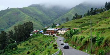 Ooty hills with winding road and tourist vehicles | JustPicked Winding mountain road through a lush green hillside village with colorful houses and misty, forested mountains in the background